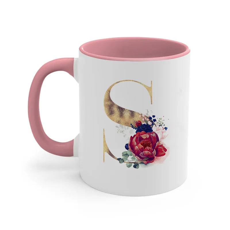 A pink ceramic coffee mug featuring a gold letter S design and floral accents.