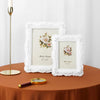 A white rose relief photo frame is displayed next to a gold candlestick and a magnifying glass on a brown table.
