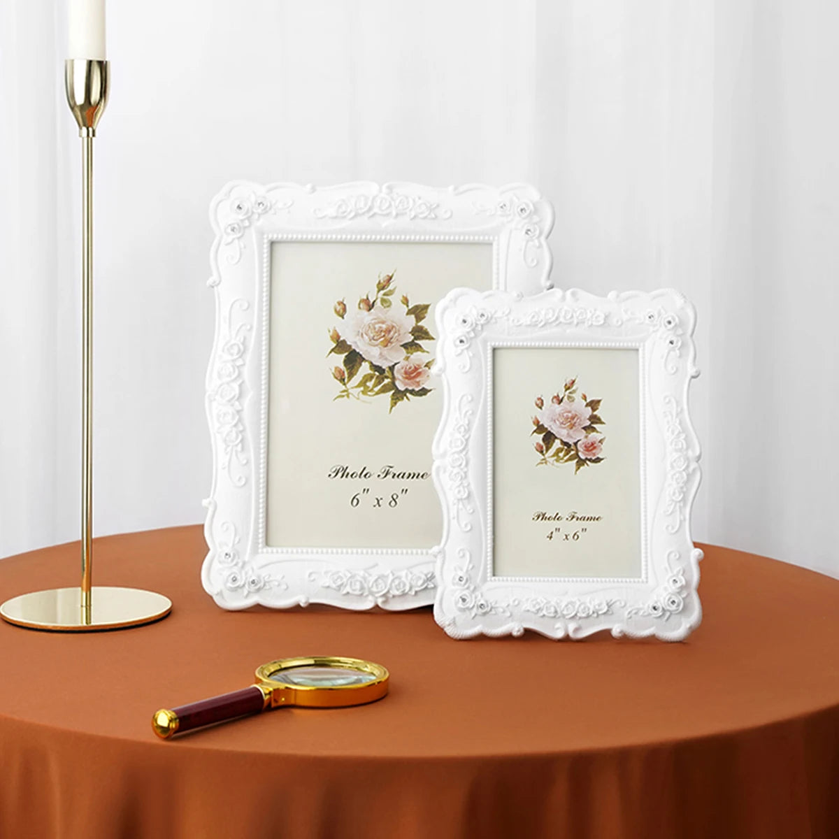 A white rose relief photo frame is displayed next to a gold candlestick and a magnifying glass on a brown table.