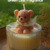 A pink teddy bear candle in a glass jar with a white ribbon set against a green outdoor background.