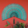 A handwoven bamboo wall fan decor in teal and pink stripes with a tassel mounted on a red background.