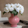 A bouquet of white artificial silk peonies in a pink vase on a wooden tray.
