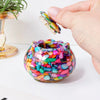A hand holds a small crystal piece above a colorful glass bowl filled with tumbled stones for meditation and home decor.
