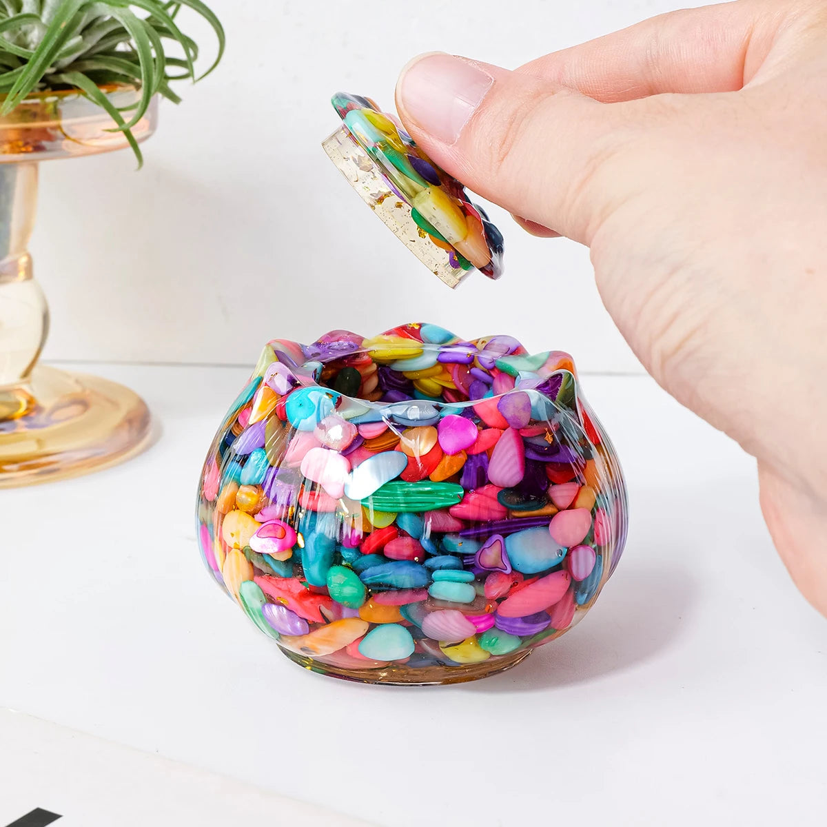 A hand holds a small crystal piece above a colorful glass bowl filled with tumbled stones for meditation and home decor.