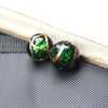 Two handcrafted lampwork glass beads with gold accents resting on a dark gray surface next to a wooden stick.