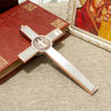 A silver crucifix wall decor featuring a St. Benedict medal and INRI inscribed on a red book, set against a beige background.