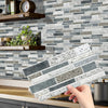 A hand is placing a rectangular crystal film marble wall decal onto a gray and white tiled backsplash.