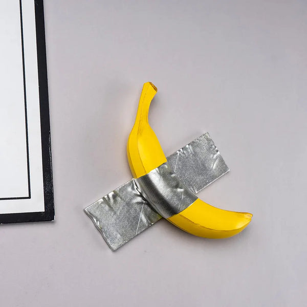 A yellow banana sculpture wrapped with silver ribbon, displayed on a light gray surface.