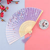 A vintage silk folding hand fan with purple and white floral patterns is displayed on a pink background.