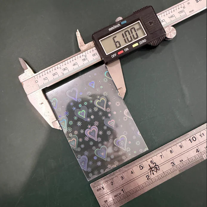 A holographic card sleeve is shown next to a digital caliper and a ruler on a green surface.