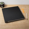 A black magnetic scrapbook album lying on a wooden surface next to an orange glue stick and a small succulent plant.