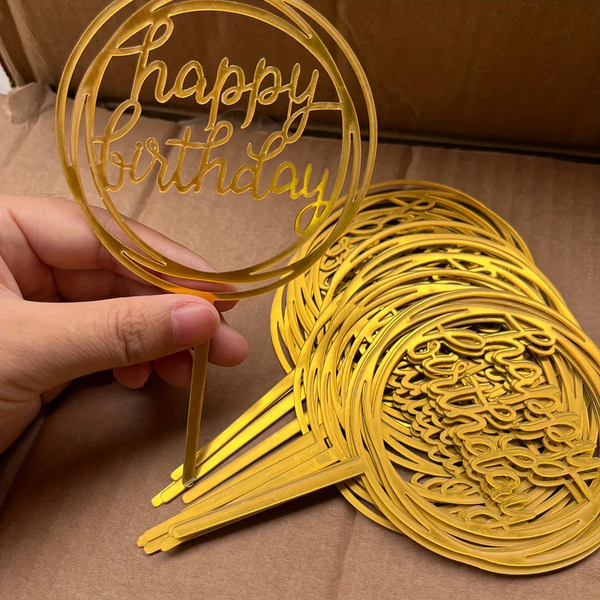 A hand holds a gold acrylic happy birthday cake topper, showing the intricate script design.