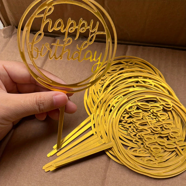 A hand holds a gold acrylic happy birthday cake topper, showing the intricate script design.