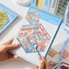 A hand holds a sticker book featuring miniature 3D scenes for kids, showing colorful scenes and stickers.