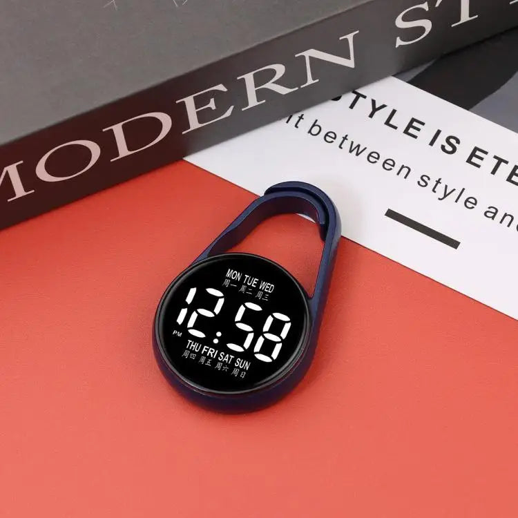 A dark blue LED pocket watch displaying 12:58 on a red surface next to a book.