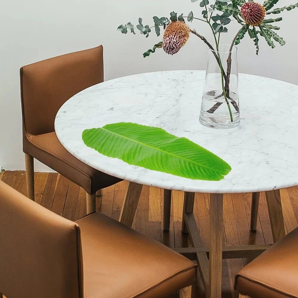 A set of five tropical banana leaf coasters displayed on a white marble table with a glass vase and wooden chairs.