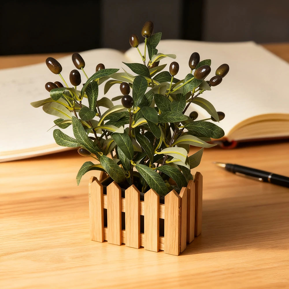 An artificial olive tree branch with green leaves and dark berries planted in a wooden box on a light wooden table.