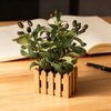 An artificial olive tree branch with green leaves and dark berries planted in a wooden planter on a light wood table.