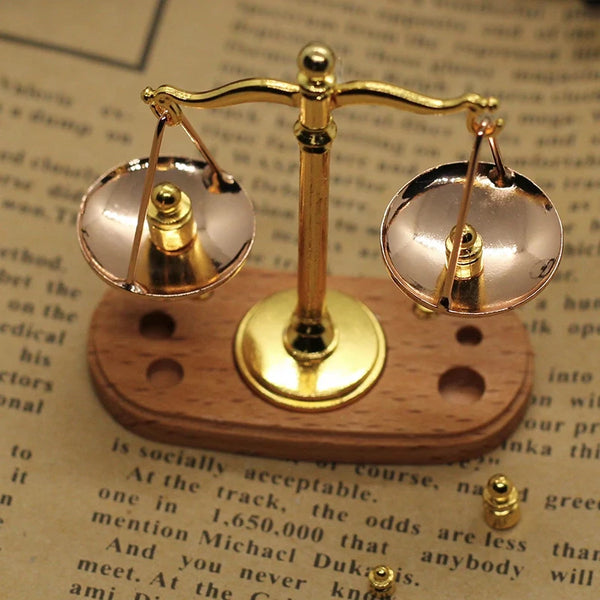 A vintage miniature balance scale with brass arms and glass pans rests on a wooden base.