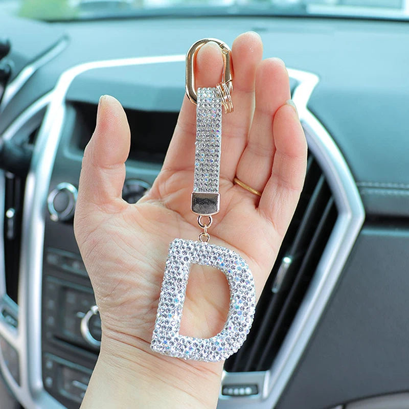 A hand holds a crystal letter keychain charm shaped like the letter D, featuring a gold-colored ring.