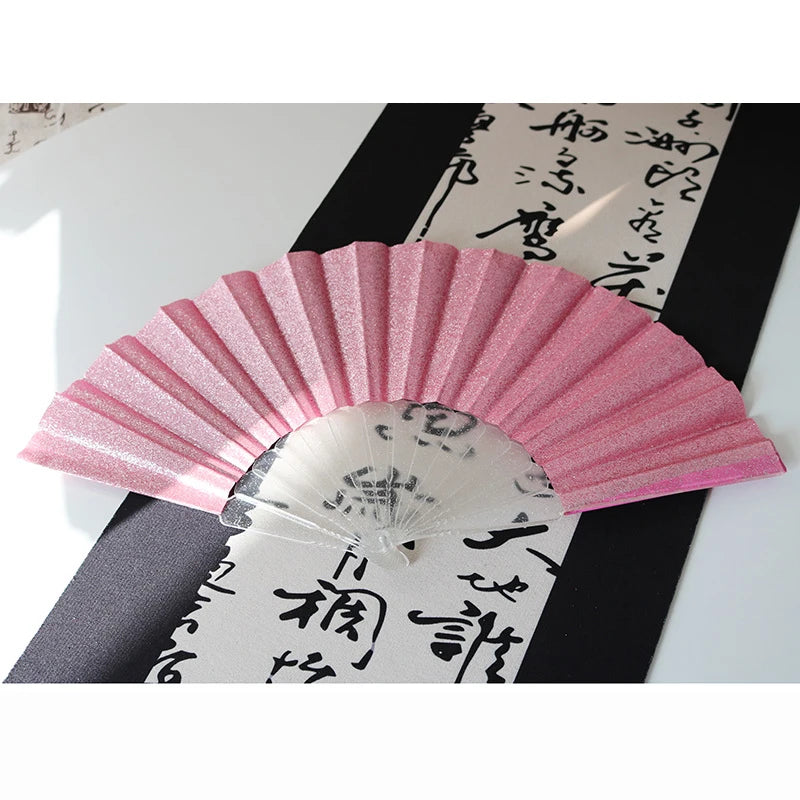 A pink glitter folding fan with transparent ribs is displayed against a black and white background.