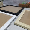 A light wood photo frame with a white border is displayed on a gray surface next to a black frame.