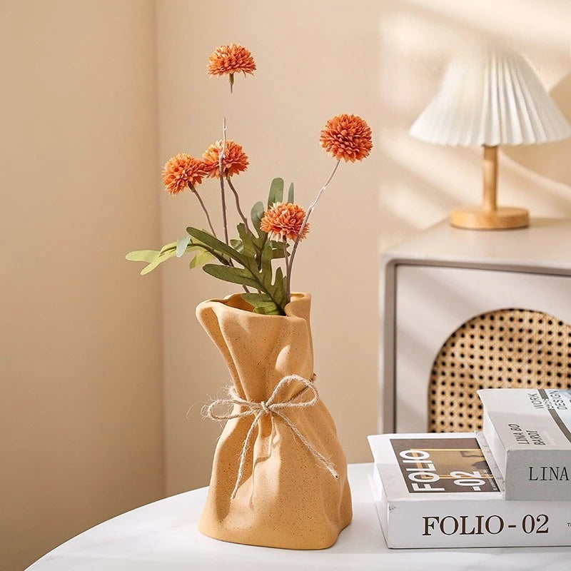A natural linen money bag shaped vase holds dried orange-red dahlia flowers on a white table.