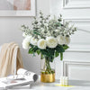 A bouquet of artificial eucalyptus stems with white flowers in a glass vase on a white surface.
