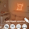 A glowing neon sign shaped like a Highland cow is mounted on a wall above a bed, with an accompanying USB power adapter visib