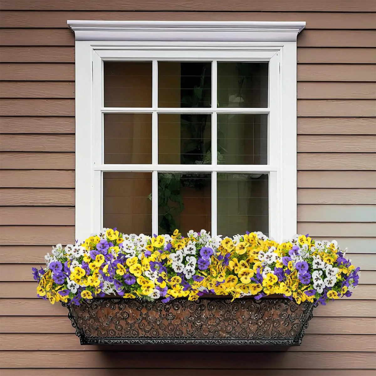 A window box filled with artificial pansy flowers sits below a white window on a brown siding wall.