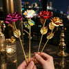 A hand holds a stem of an iridescent gold foil rose with vibrant pink and gold petals against a city skyline backdrop.