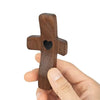A hand holds a dark brown wooden cross with an epoxy heart carved into the center.