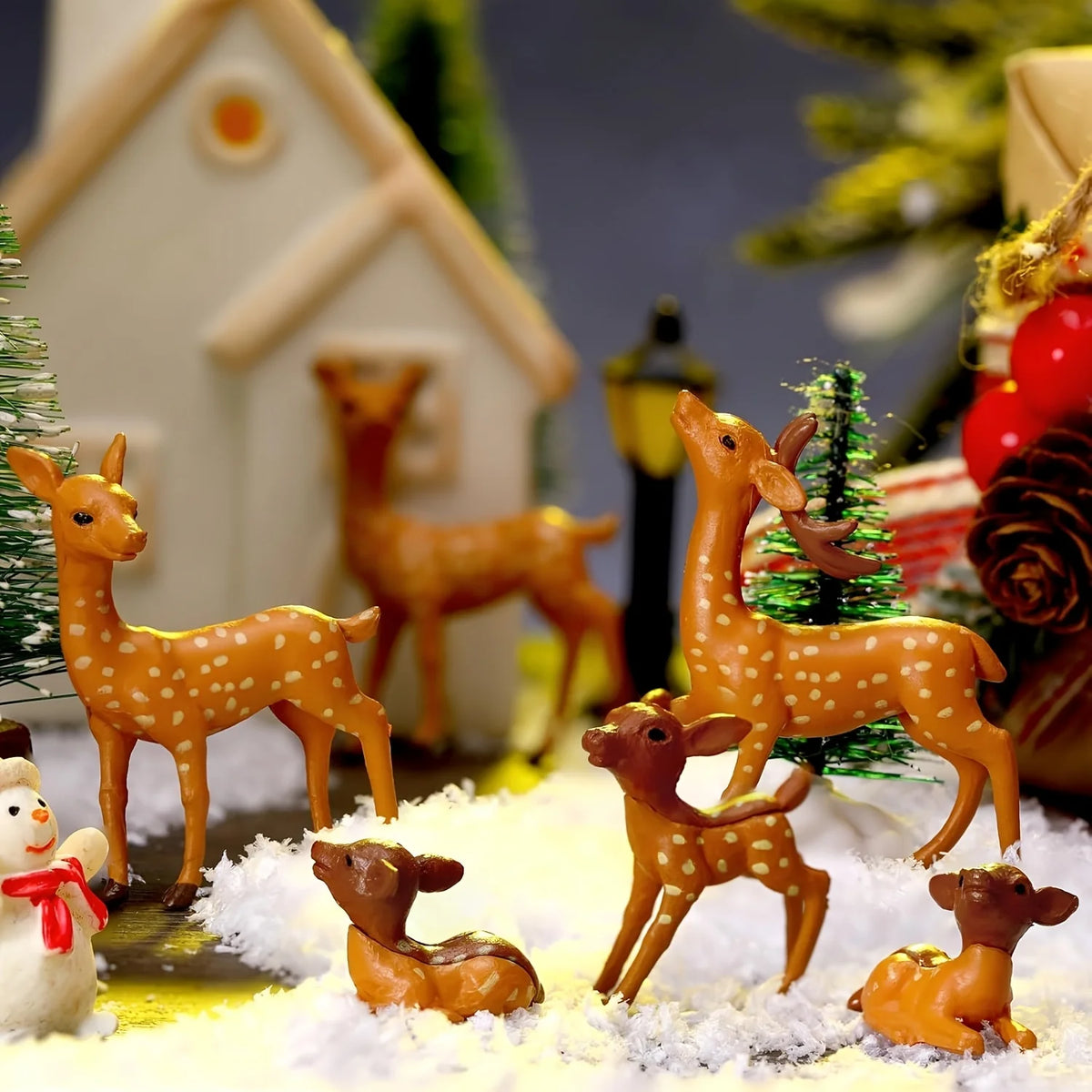 A set of small orange deer figurines standing on white snow, set against a backdrop of a white house and green foliage.