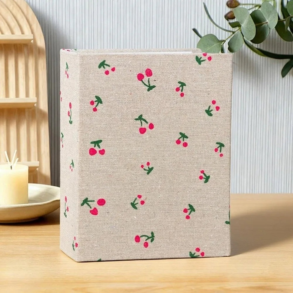 Beige Leinen-Cover-Fotoalbum mit wiederkehrendem Muster aus rosa und grünen Kirschen auf einer Holzoberfläche.