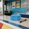 A roll of blue tearable raffle tickets sits on a counter in an office with colorful framed posters in the background.