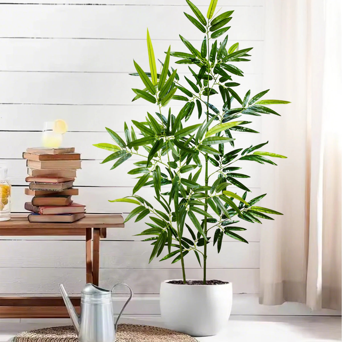 An artificial bamboo branch with green leaves stands in a white pot next to a stack of books and a metal teapot.