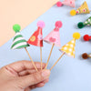 A hand holds a set of mini birthday cake toppers featuring colorful conical shapes and fuzzy pom-poms.