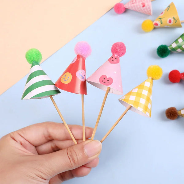 A hand holds a set of mini birthday cake toppers featuring colorful conical shapes and fuzzy pom-poms.