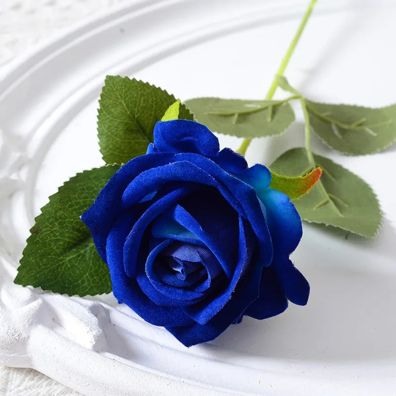 A deep blue velvet artificial rose with green leaves on a white plate.