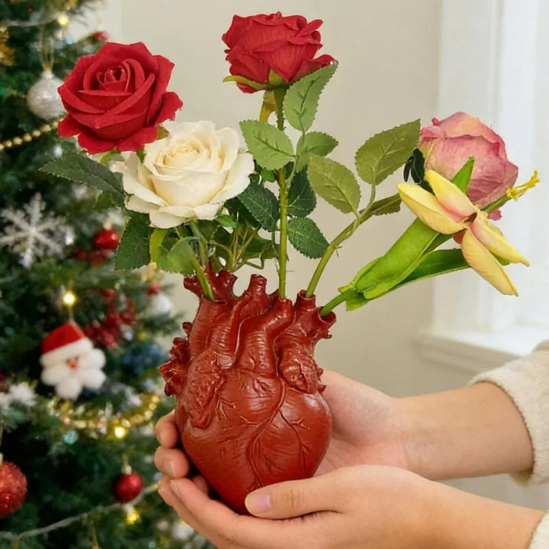 Hands hold a heart-shaped resin vase filled with red and cream roses next to a decorated Christmas tree.