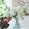 A bouquet of large white artificial hydrangea flowers with green leaves displayed in a clear glass vase.