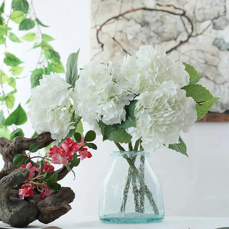 A bouquet of large white artificial hydrangea flowers with green leaves displayed in a clear glass vase.