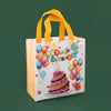 A white gift bag with yellow handles decorated with colorful balloons and a birthday cake design.