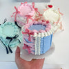 A hand holds a realistic 4-inch decorative fake cake with pink and blue frosting and ribbons on a clear plate.