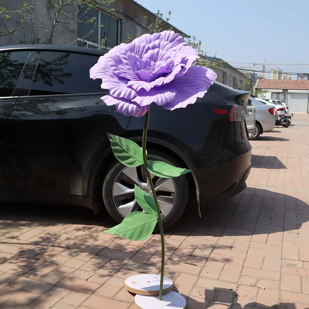 A purple artificial peony flower with green leaves is displayed in front of a black car.