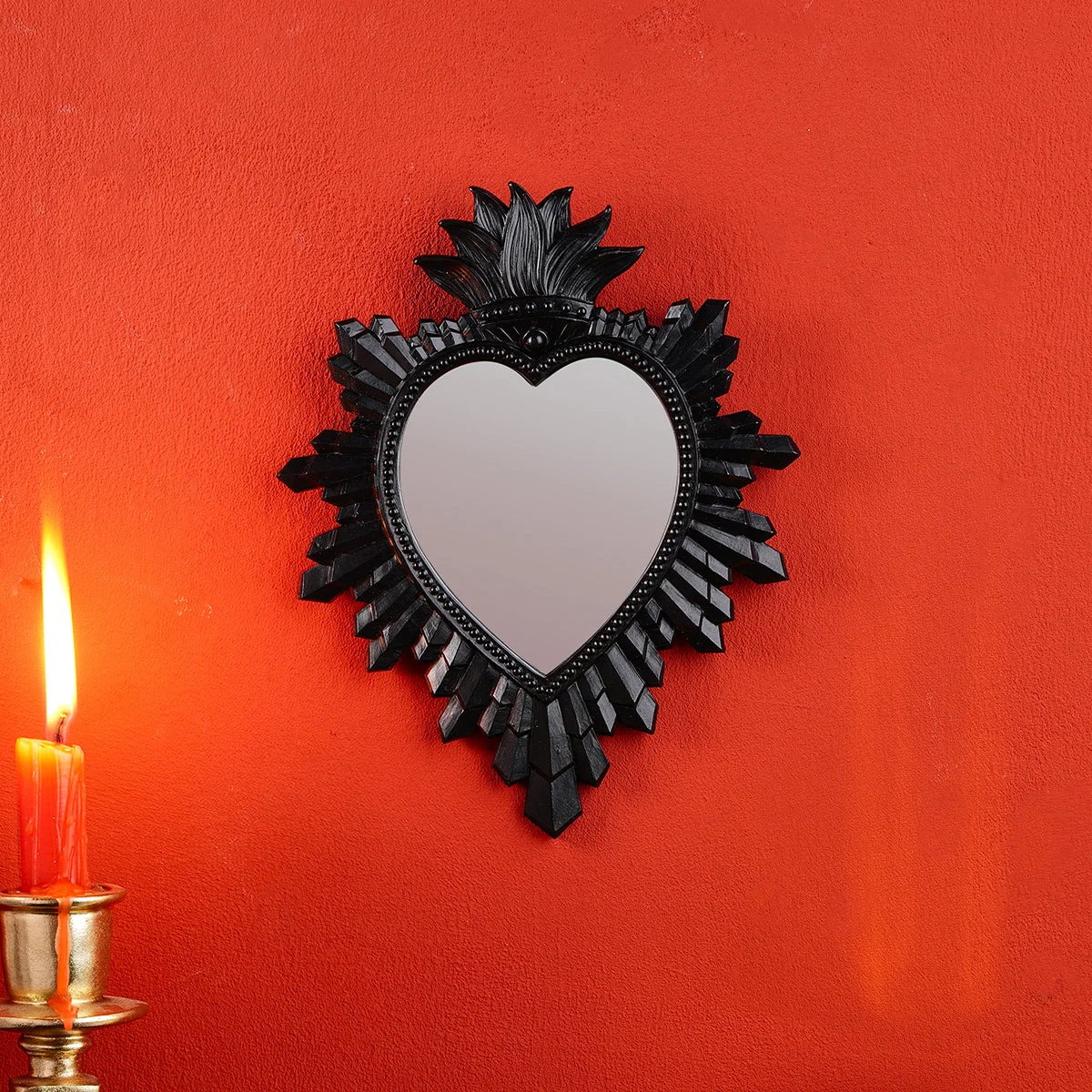 A black heart-shaped wall pendant with a mirror finish is displayed on an orange wall next to a lit candle.