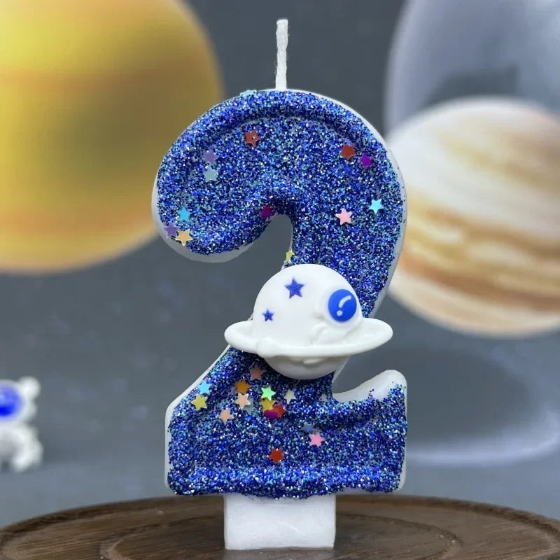 A blue glitter candle shaped like the number 2 featuring a galaxy design and a white planet on a wooden surface.