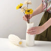 A hand holds a white plastic vase with yellow and cream flowers on a white table.