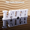 White PVC table ornament spelling out the word LAUGH on a reflective surface.