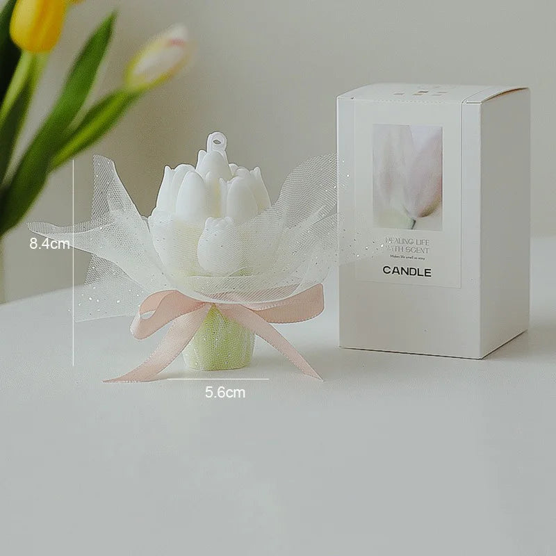 A white candle in a box with a floral design and a silk ribbon, set against a background of yellow tulips.